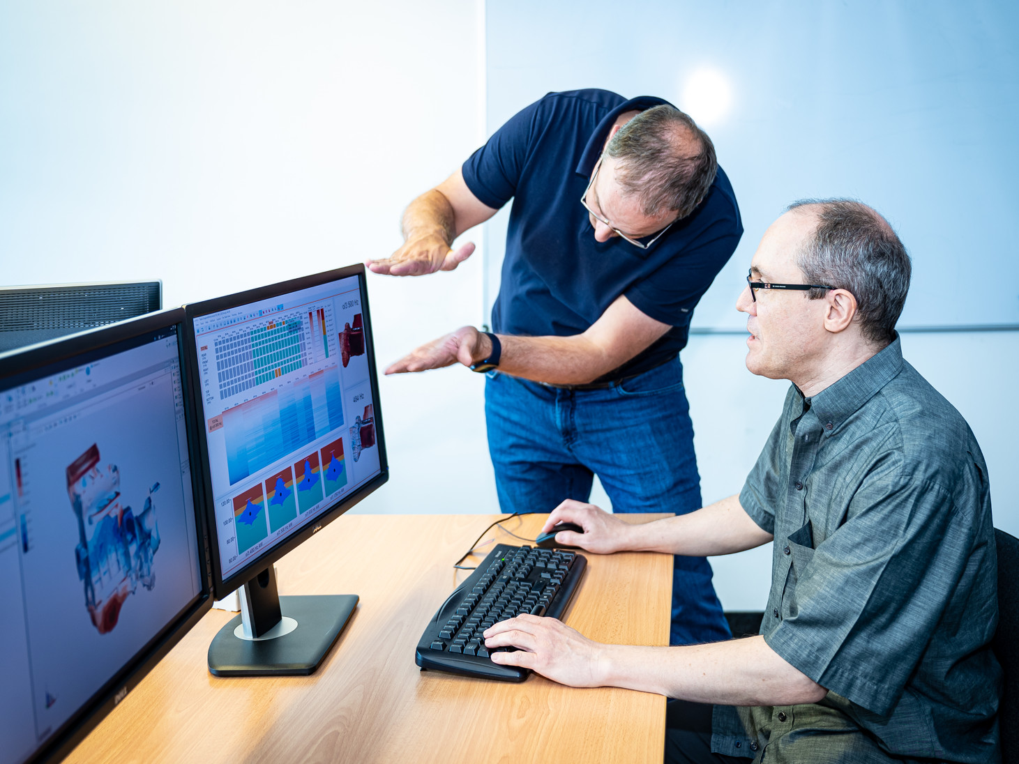 Zwei Forscher besprechen Geräuschsimulations-Visualisierungen aus der Motorenentwicklung, die auf zwei Bildschirmen dargestellt werden. 