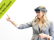 Eine Forscherin mit Augmented Reality-Brille deutet mit dem Finger nach links oben, wo sich in der Bildecke ein grünes Banner mit dem Text "Nominiert für Women in Tech-Award" befindet.