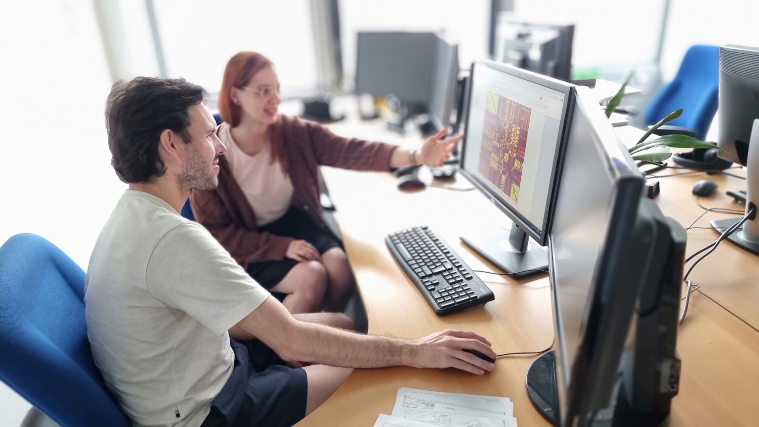 Eine Frau und ein Mann sitzen nebeneinander vor zwei Computerbildschirmen, die Frau zeigt auf einen der Bildschirme