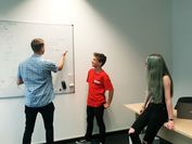 Ein Forscher schreibt auf ein Whiteboard, während zwei Schülerpraktikanten interessiert zuhören.