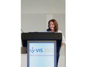 A woman is standing behind a lectern, which includes a screen showing the title of the event "VIS 2023"Titel einer Konferenz