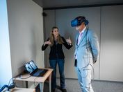 A man in a suit with VR glasses, next to him a woman gesturing with her hands to explain something. 