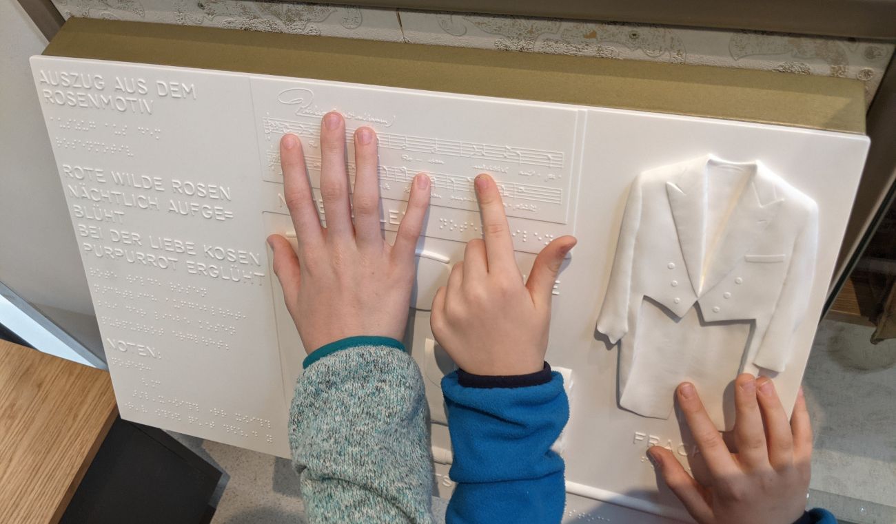 Drei Hände ertasten ein taktiles Relief, das unter anderem einen Frack und Notenzeilen zeigt