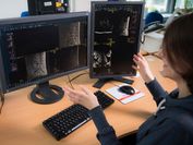 A researcher points towards two computer screens on which you can see the semnatic annotation of a human spine.Eine Forscherin zeigt auf zwei Bildschirme, wo annotierte Wirbelsäulen zu sehen sind.