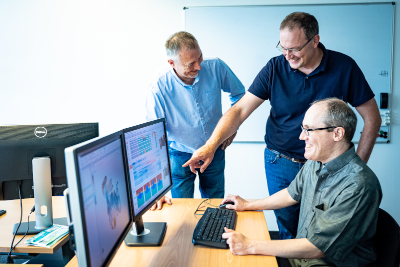 Ein Forscher sitzt vor einem Desktop-Computer mit zwei Bildschirmen auf denen bunte Datenvisualisierungen zu sehen sind, zwei weitere Forscher des VRVis stehen daneben. Ein Forscher zeigt auf den Bildschirm.
