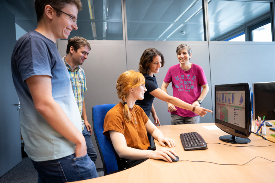 Eine Forscherin sitzt an einem Desktop-Computer, umringt von ihren Kolleginnen und Kollegen. Eine Forscherin zeigt auf den Bildschirm, auf welchem verschiedene bunte Datenvisualisierungen zu sehen sind.
