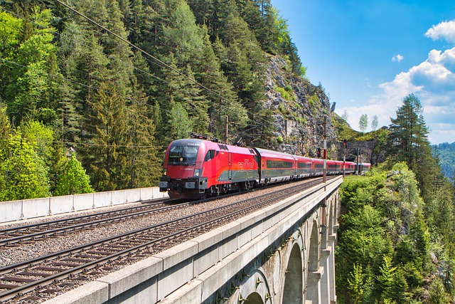 A red ÖBB train crosses a bridge, with a wooded mountain landscape in the background.
