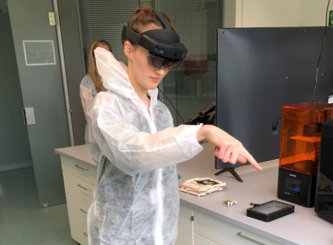 A researcher in a lab suit stands in a lab with her hand in the air while using an augmented reality application.