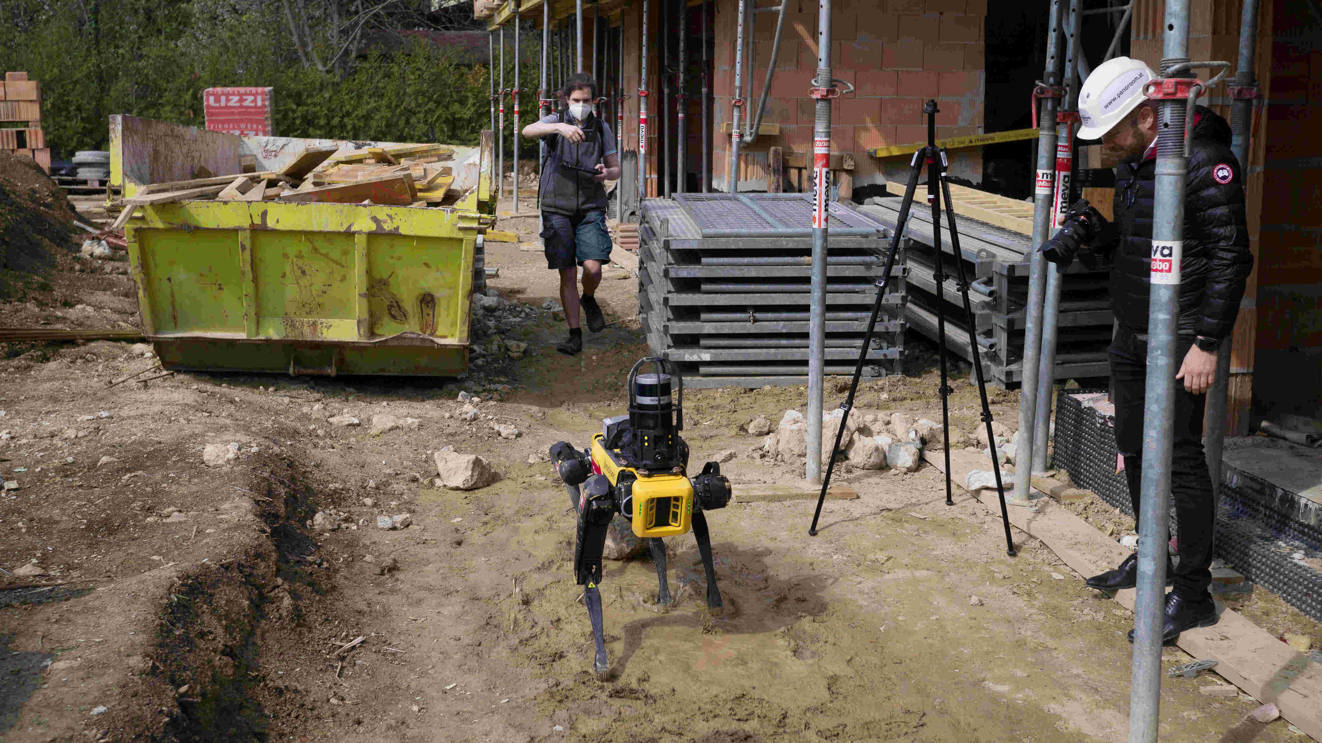 Der Laufroboter Spot von vorne, inmitten einer Baustellenszene, inklusive Rohbau und Mulde. 