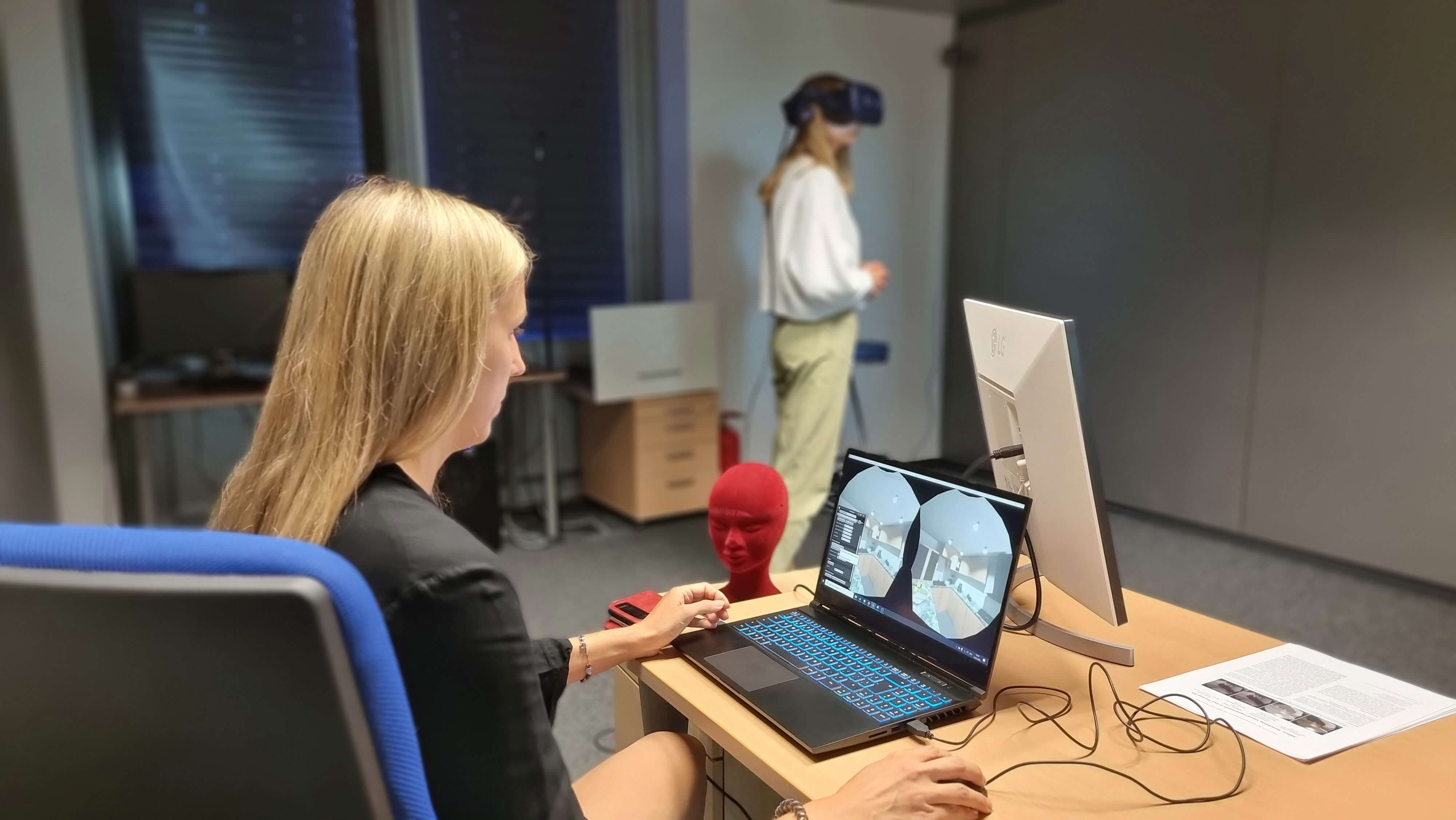 A woman sits at a laptop, behind her a second woman moves around the room standing with VR glasses. 