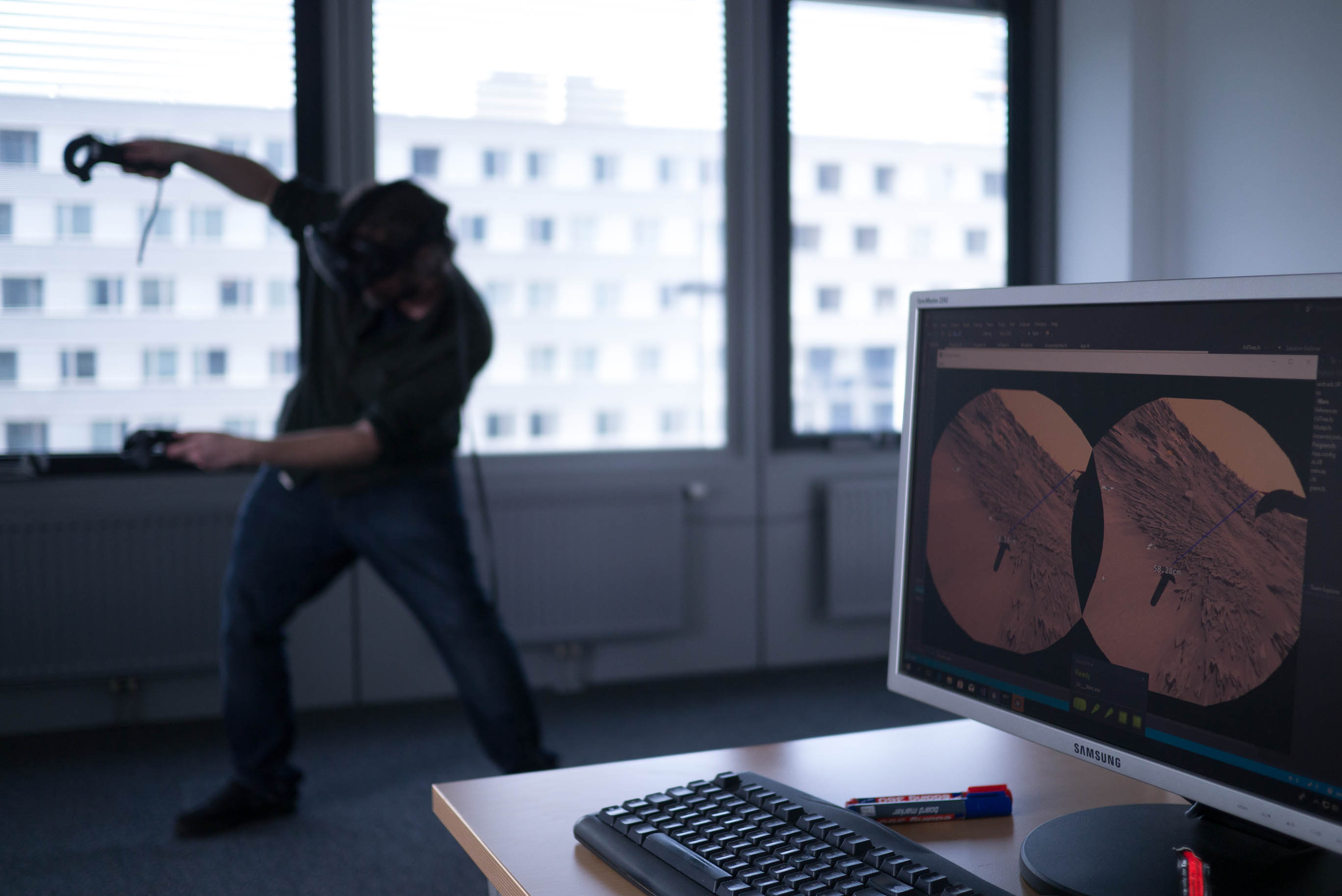 In the foreground is a desktop computer where 3D reconstructions of the Martian surface can be seen. In the background is a man with virtual reality gear moving around in this data in VR and exploring the Martian surface.