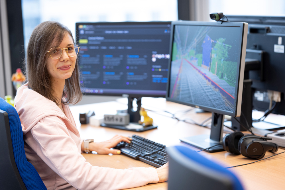Eine Frau sitzt vor einem Computer mit zwei Bildschirmen, auf dem linken Bildschirm ist eine Software-Library zu sehen, auf dem anderen Bildschirm ist eine Bahnsteig-Rekonstruktion aus einer Punktwolke zu sehen.
