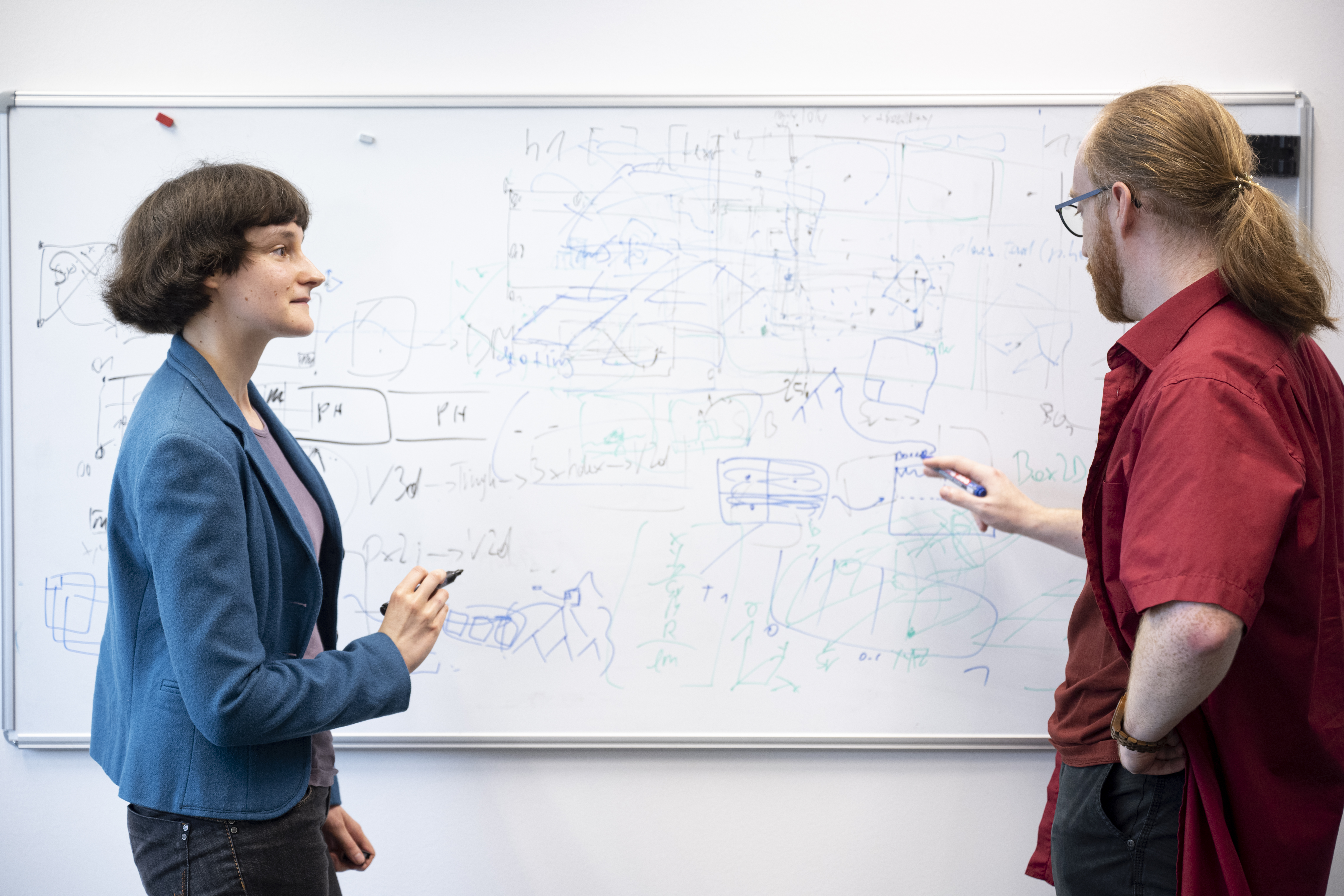 Eine Forscherin (links) und ein Forscher (rechts) stehen vor einem dicht beschriebenen Whiteboard und diskutieren eine Forschungsfrage.