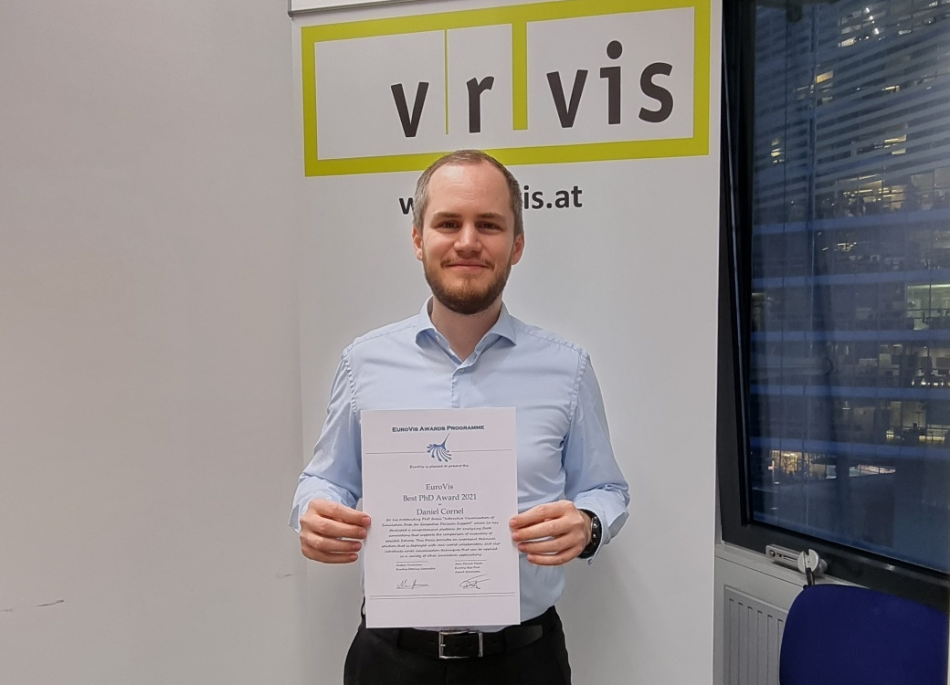 A man stands in front of a VRVis banner and holds his award certificate into the camera.