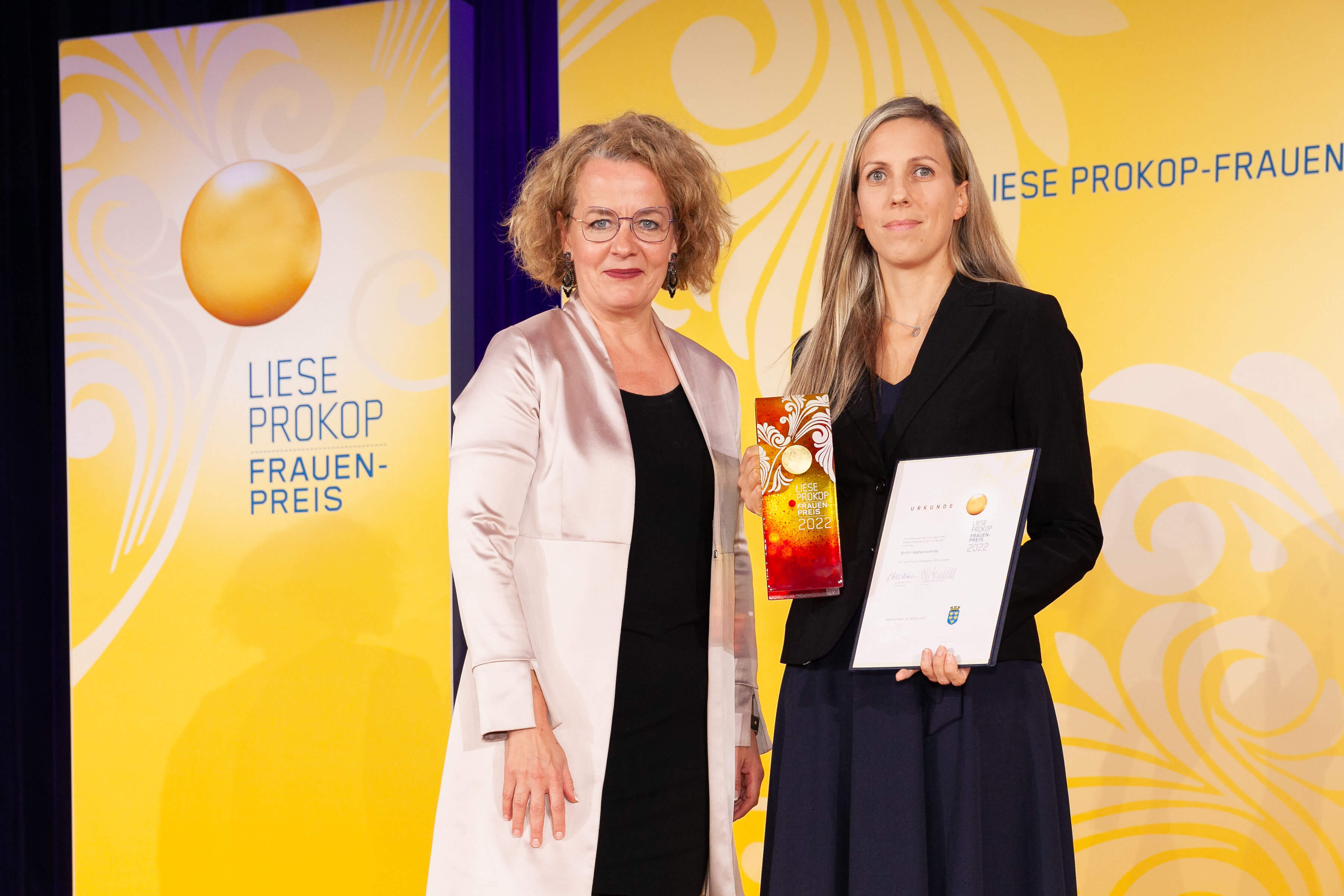 Das Logo des Liese-Prokop-Frauenpreises auf gelbem Hintergrund links, rechts daneben stehen zwei Frauen.