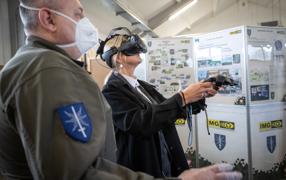 Eine Frau hat eine VR-Brille aufgesetzt und bewegt sich mithilfe der VR-Controller in der virtuellen Realität, ein Bundesheer-Soldat steht an ihrer Seite.