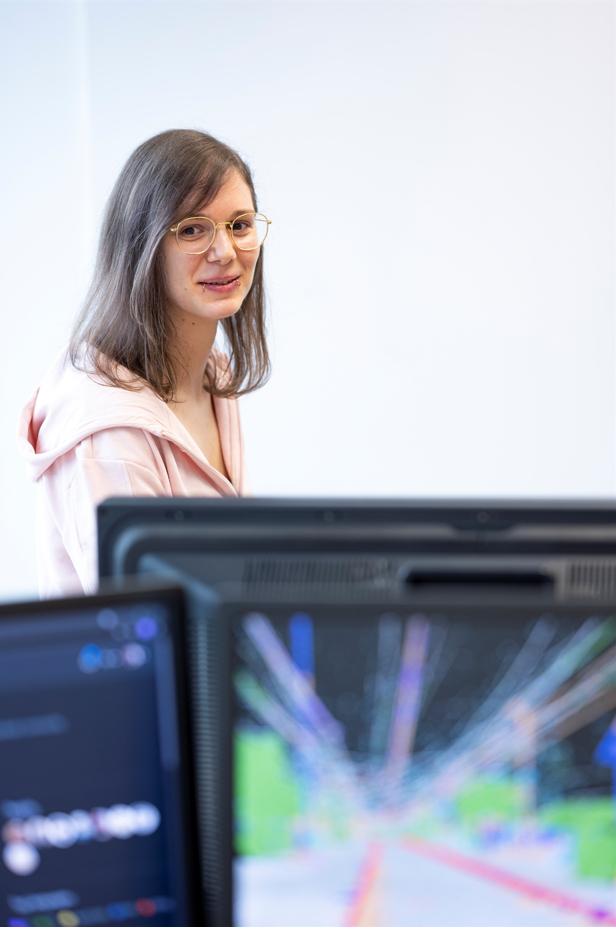 Eine Forscherin steht hinter Bildschirmen, auf denen Bahnsteig-Grafiken leuchten. 
