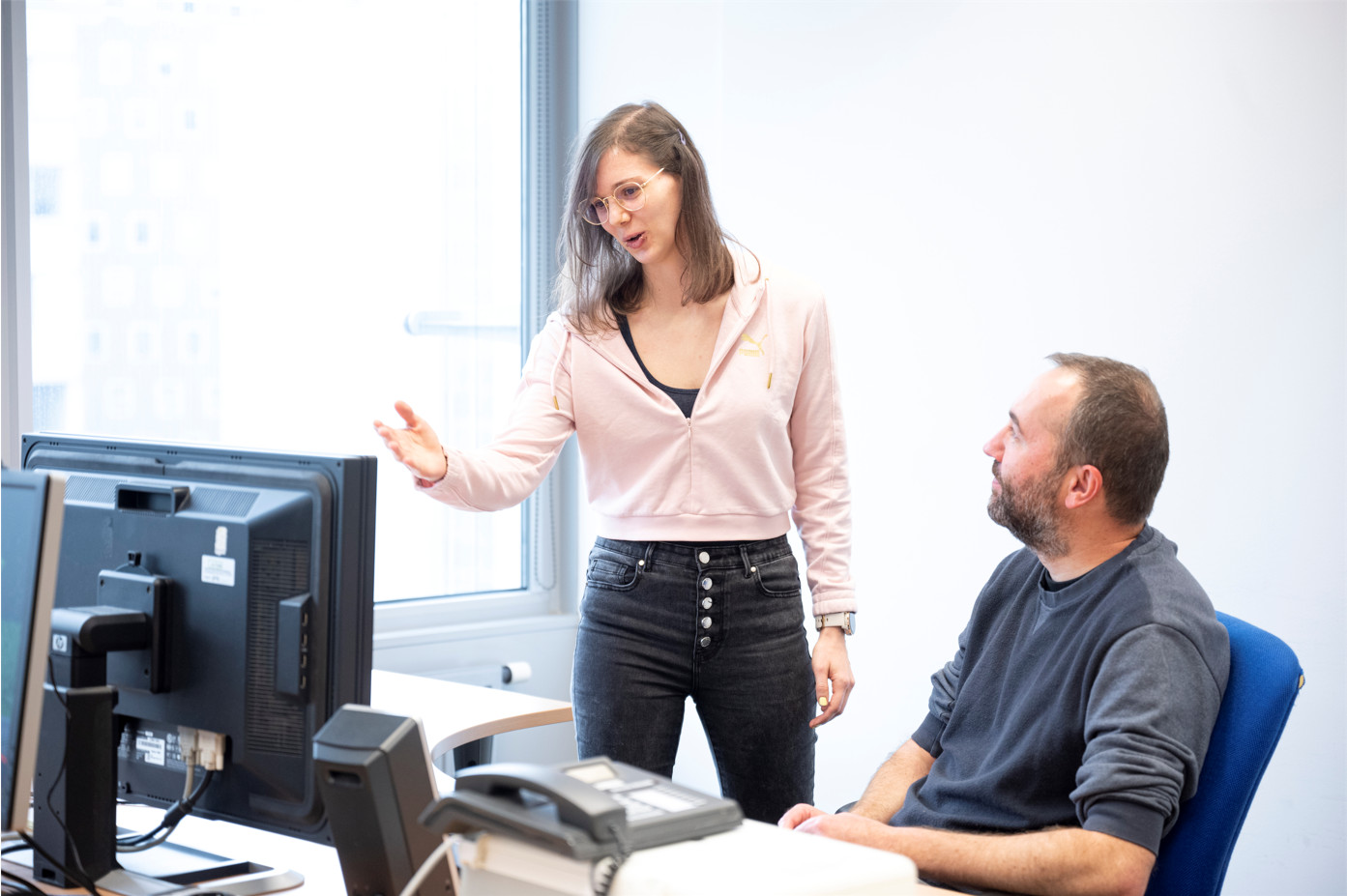 A researcher, on the left, standing, explains something on the screen to a researcher, on the right, sitting.