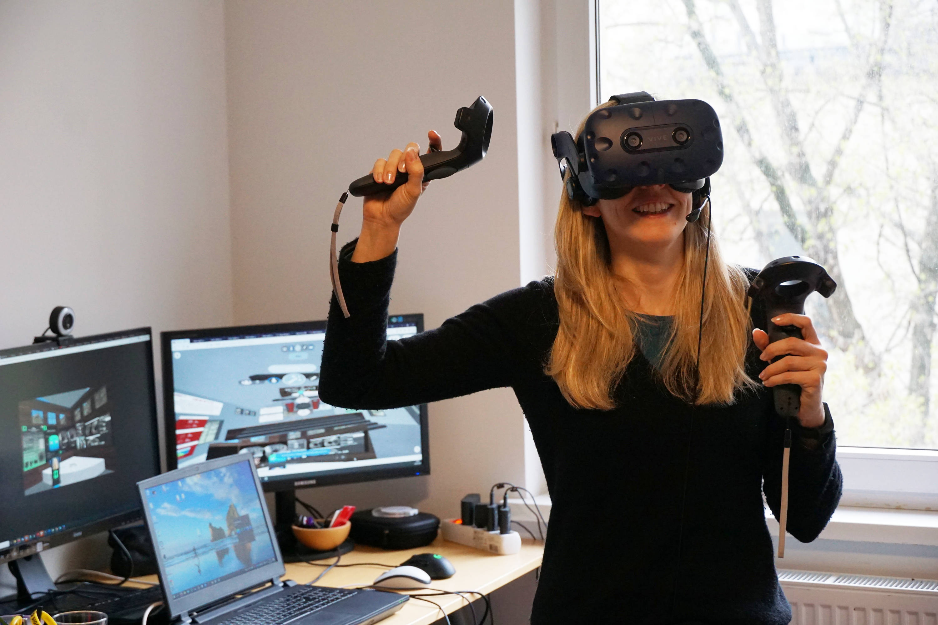 A researcher with VR goggles and controllers in front of three computer monitors. 