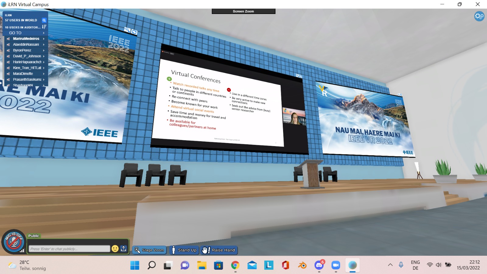 Screenshot eines virtuellen Konferenzraums mit Bühne und einer Wand voller Präsentationsflächen