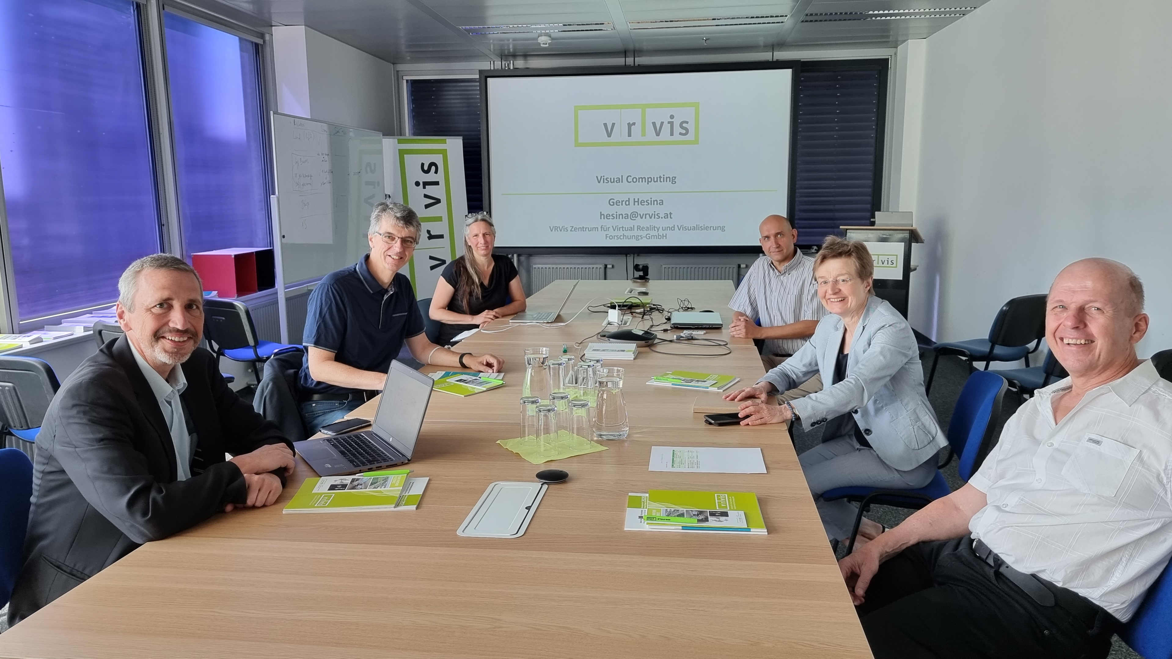 Eine Gruppe von Forscherinnen und Forscher an einem langen Tisch. Am Ende ist ein Bildschirm mit dem VRVis-Logo zu sehen.