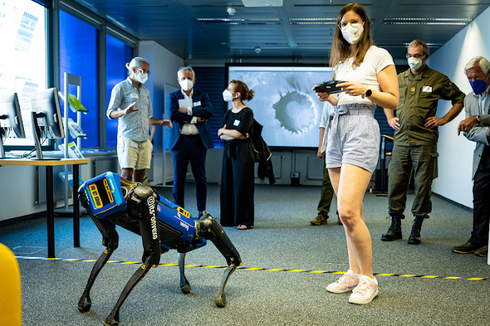 A robot dog and a researcher with MNS stand in a large meeting room, while in the background several guests also with MNS stand and watch.