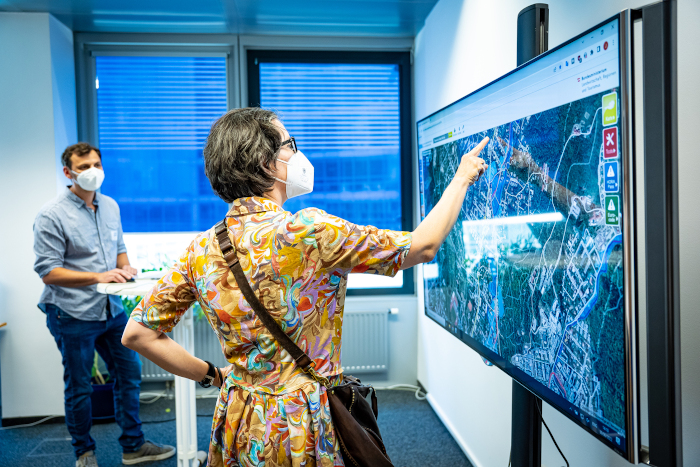 Eine Frau mit MNS steht an einem großen Bildschirm und deutet auf einen Punkt auf der Hochwasserrisikolandkarte Österreichs, im Hintergrund steht ein Forscher des VRVis ebenfalls mit MNS und erklärt ihr, was sie auf dem Bildschirm genau sehen kann.