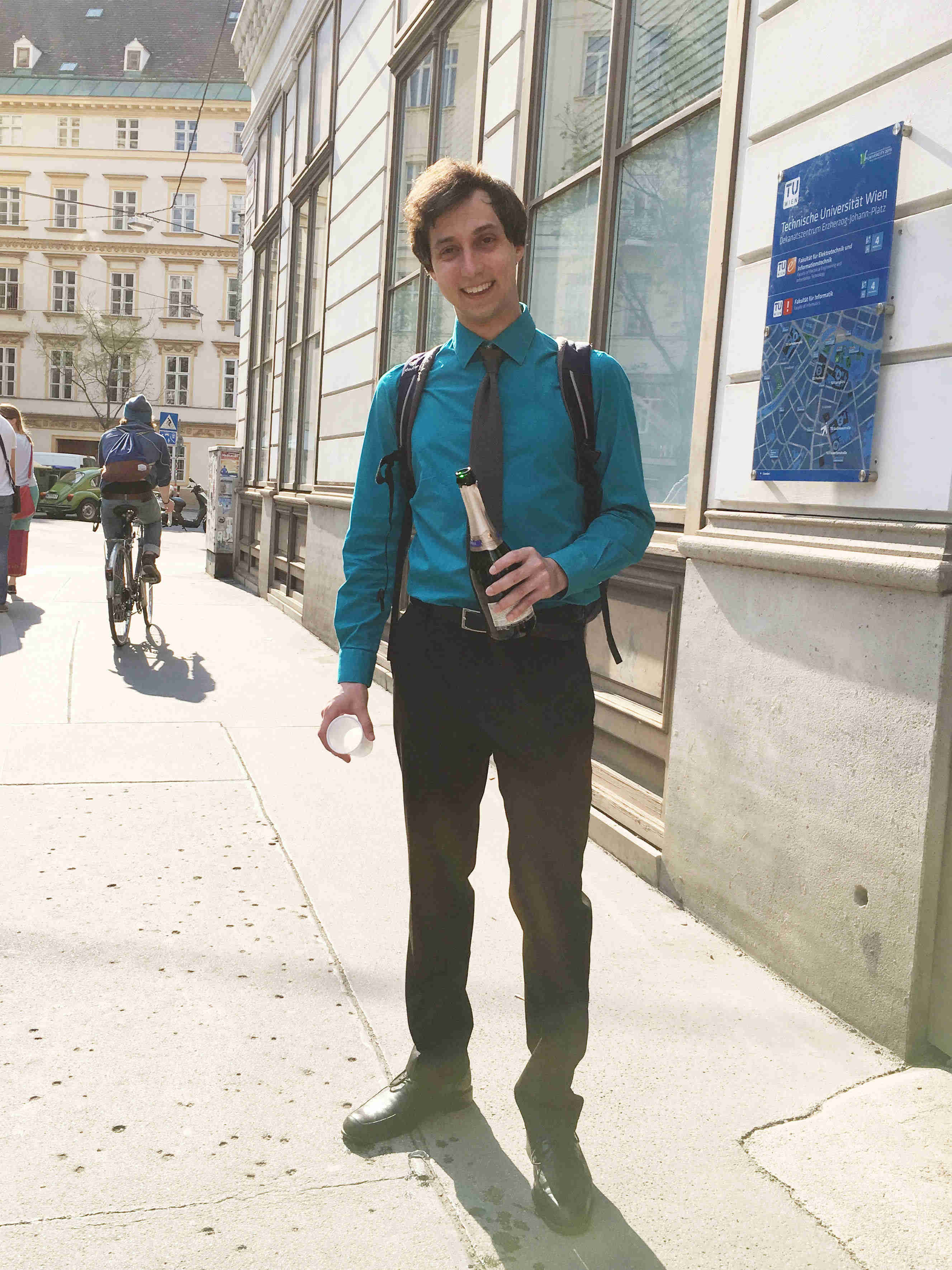Image of a researcher in shirt and tie holding a champagne bottle in front of a university building.
