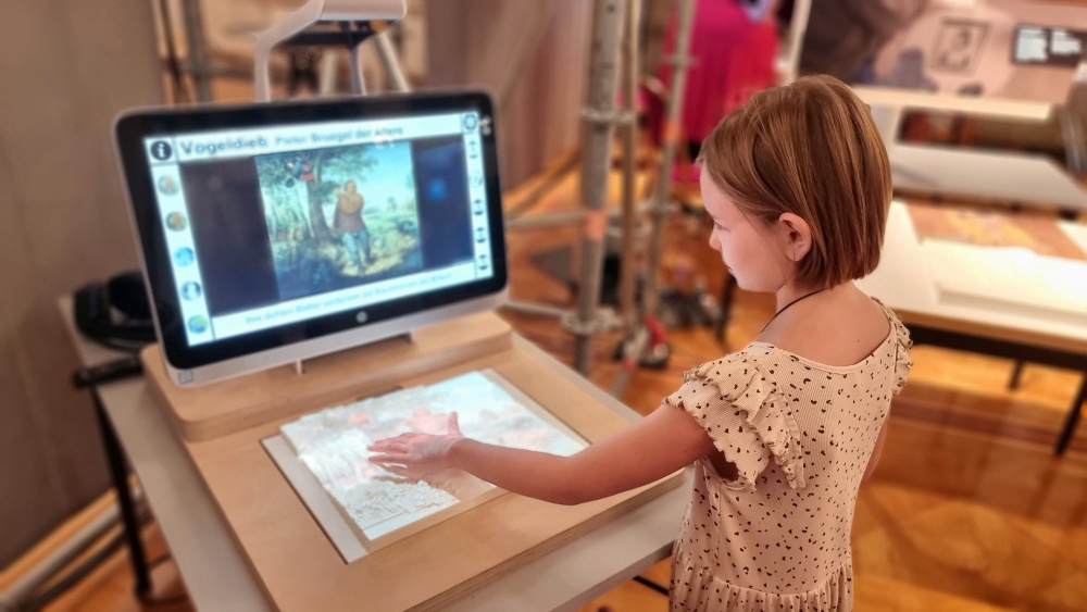 A girl touches a tactile relief of Pieter Breughel's Bird Thief in front of a screen showing a digital version of the painting.