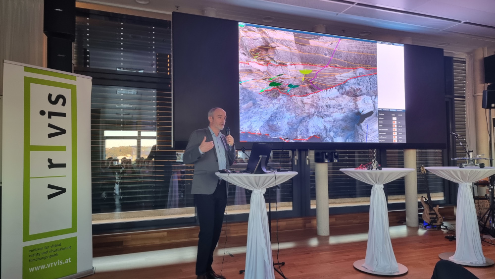 A man has a microphone in his hand and speaks to the audience, in the background hangs a large screen on which geological annotations on Mars can be seen. To the left is the VRVis banner.