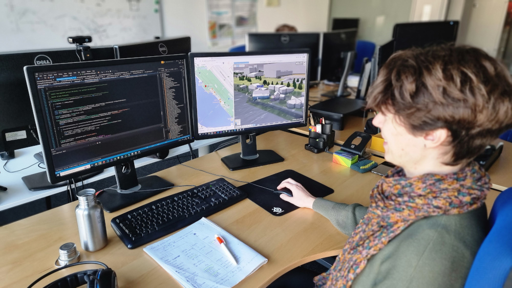 A researcher sits at her workstation in front of two screens; the left screen shows lines of programming code, while the right screen shows a flood visualization for the city of Vienna from VRVis.