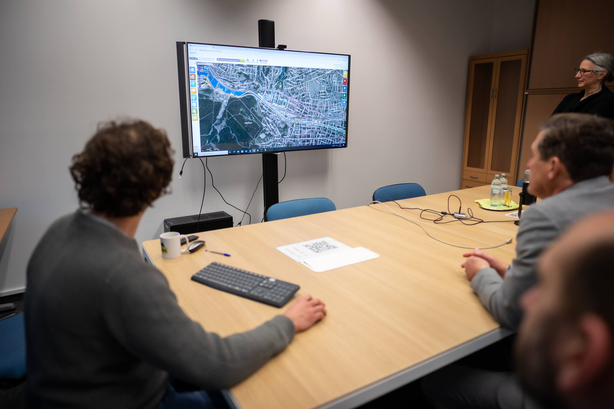Menschen schauen auf einen großen Monitor, auf dem eine 3D-Darstellung eines Stadtviertels zu sehen ist. 