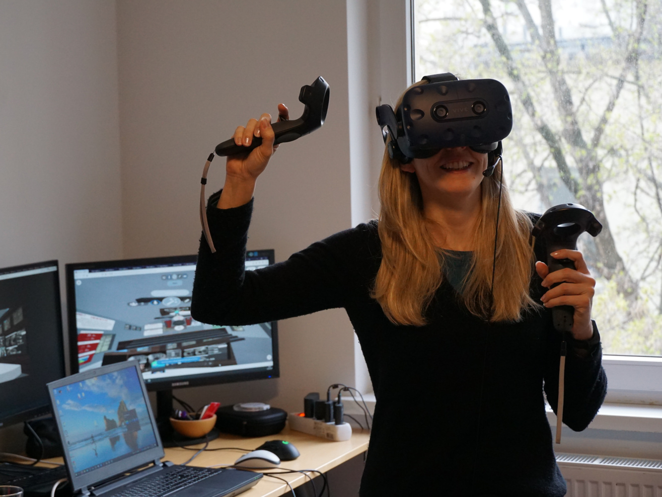 Katharina Krösl works in her home office: she has a VR headset on and controllers in her hand; to her left are several computer screens.