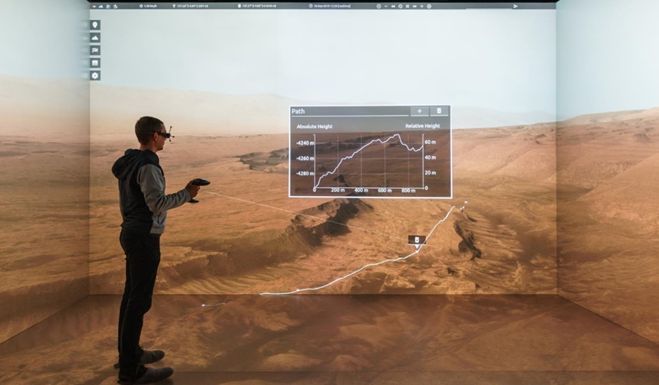 Ein Mann mit Virtual Reality Hardware steht in einer dreiseitigen Cave-Umgebung auf welche projiziert wird, was er in VR sieht. Es handelt sich um eine 3D-Rekonstruktion einer Planetenoberfläche.