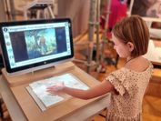 A girl touches a tactile relief of Pieter Breughel's Bird Thief in front of a screen showing a digital version of the painting.