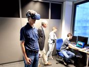 Im Vordergrund steht ein Mann, der eine VR-Brille trägt, im Hintergrund schauen zwei Personen einem Forscher über die Schulter, der am Computer die VR-Anwendung bedient.