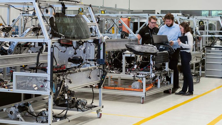 Drei Menschen stehen hinter einem Laptop in einer Fabrikhalle, umgeben von Regalen mit Kabeln und anderen technischen Gegenständen
