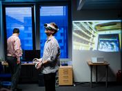 Ein Mann trägt eine VR-Brille und steht mitten in einem Raum, im Hintergrund ist eine große Projektion, die zeigt, was der Mann in Virtual Reality sieht.