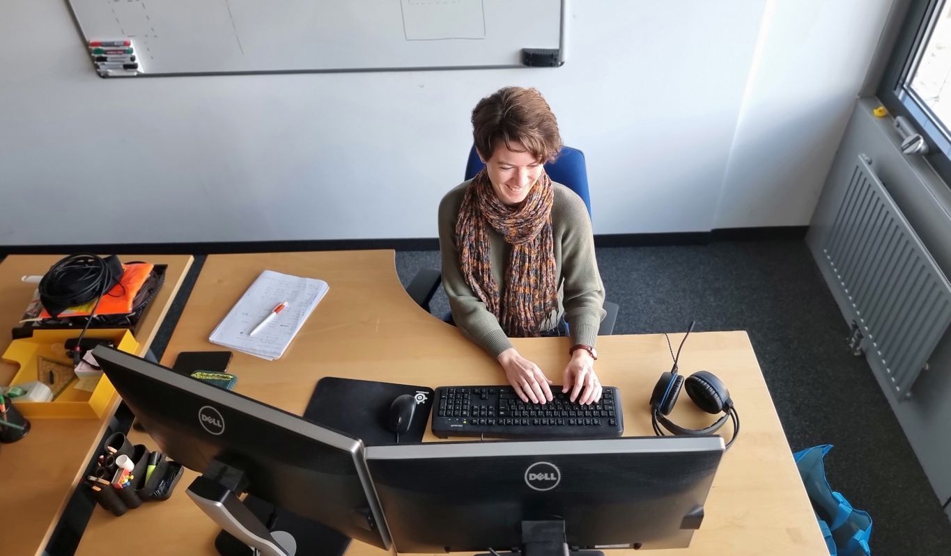 Sicht auf eine Forscherin, die an ihrem Arbeitsplatz vor einem Computer mit zwei Bildschirmen sitzt.