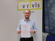 A man stands in front of a VRVis banner and holds his award certificate into the camera.