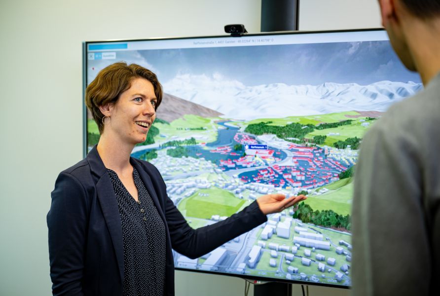 VRVis-Forscher Silvana Rauer-Zechmeister zeigt auf einen großen Bildschirm, auf welchem eine Hochwassersimulation zu sehen ist.