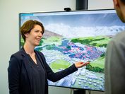 VRVis-Forscher Silvana Rauer-Zechmeister zeigt auf einen großen Bildschirm, auf welchem eine Hochwassersimulation zu sehen ist.