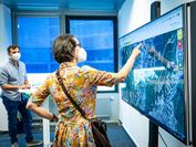 Eine Frau mit MNS steht an einem großen Bildschirm und deutet auf einen Punkt auf der Hochwasserrisikolandkarte Österreichs, im Hintergrund steht ein Forscher des VRVis ebenfalls mit MNS und erklärt ihr, was sie auf dem Bildschirm genau sehen kann.
