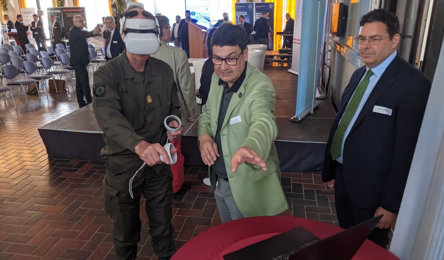 A man is wearing VR goggles, next to him is a second man, pointing at a computer screen. Another man is standing next to them.
