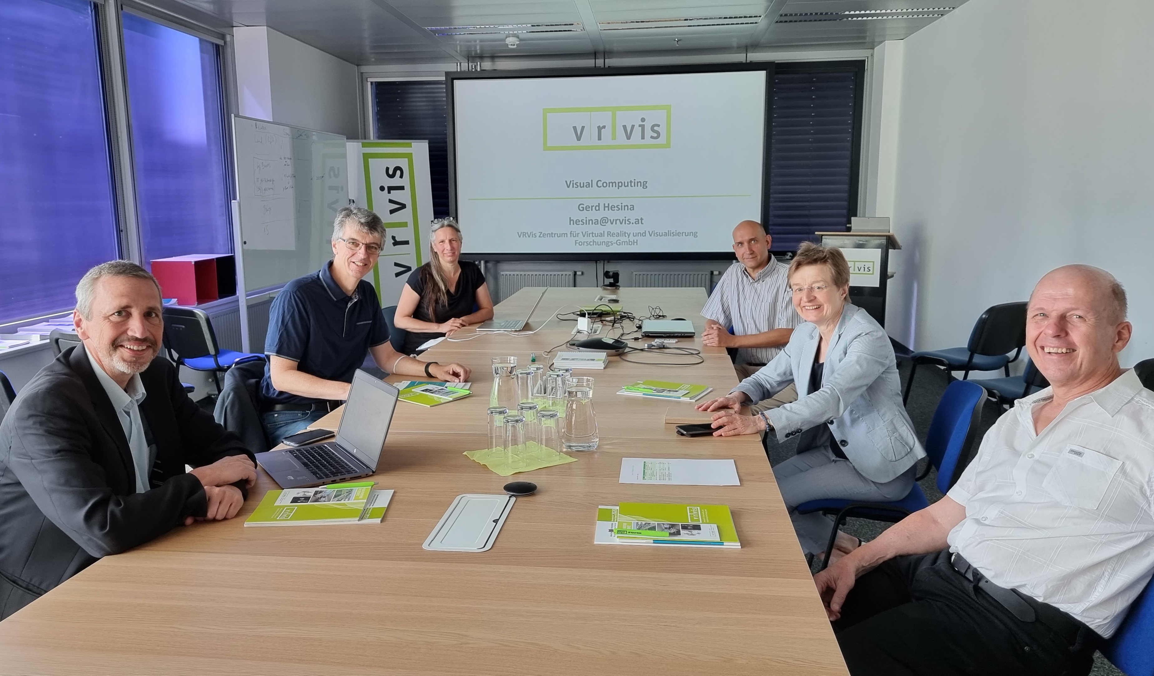 Eine Gruppe von Forscherinnen und Forscher an einem langen Tisch. Am Ende ist ein Bildschirm mit dem VRVis-Logo zu sehen.