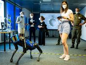 A robot dog and a researcher with MNS stand in a large meeting room, while in the background several guests also with MNS stand and watch.