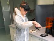 A researcher in a lab suit stands in a lab with her hand in the air while using an augmented reality application.
