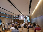 Bild von einem Konferenzraum mit Menschen und einem großen Vortragsbildschirm. 
