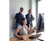 Eine junge Forscherin sitzt mit Mund-Nasen-Schutz am Computer und zeigt zwei hinter ihr stehenden Männern eine Punktwolken-Demo am Computer. 