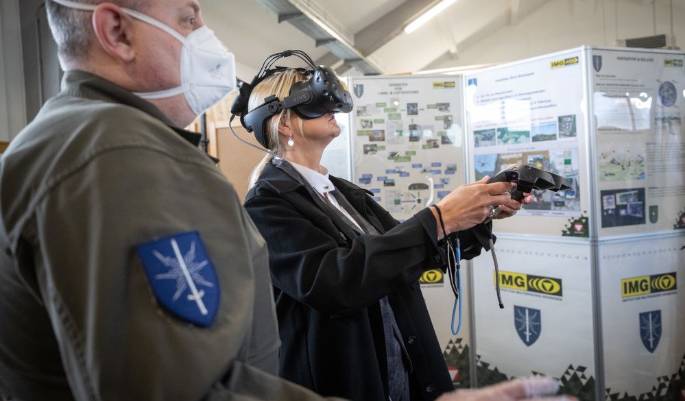 Eine Frau hat eine VR-Brille aufgesetzt und bewegt sich mithilfe der VR-Controller in der virtuellen Realität, ein Bundesheer-Soldat steht an ihrer Seite.
